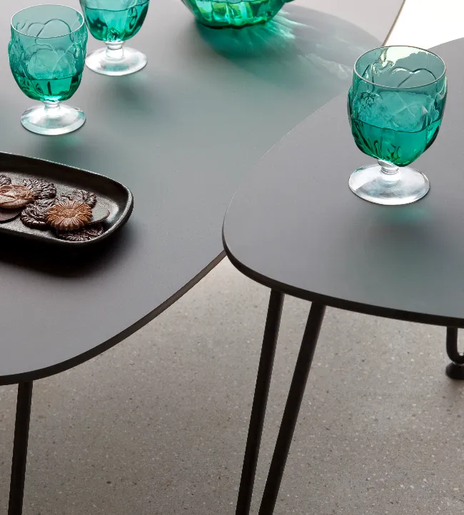 Close-up of Rozy tables by Vincent Sheppard with smooth grey HPL tops and black metal hairpin legs, featuring green textured glassware and a black ceramic tray.