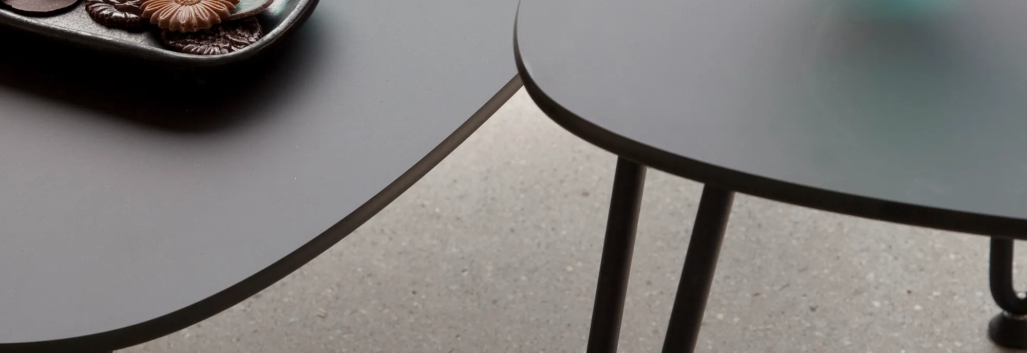 Close-up of Rozy tables by Vincent Sheppard with smooth grey HPL tops and black metal hairpin legs, featuring green textured glassware and a black ceramic tray.