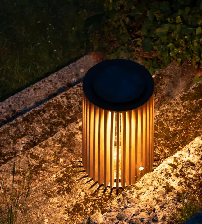 Maya solar lantern by Vincent Sheppard, made of vertical teak slats with a black top, softly glowing with warm light in an outdoor setting.