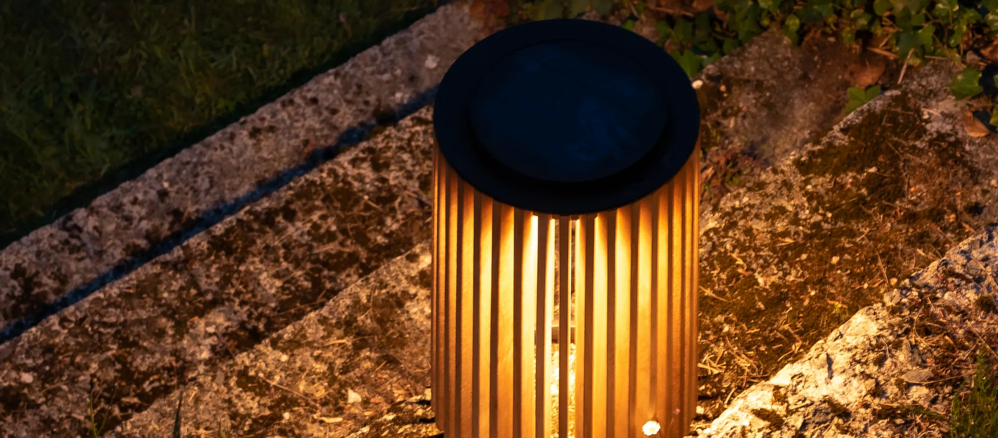 Maya solar lantern by Vincent Sheppard, made of vertical teak slats with a black top, softly glowing with warm light in an outdoor setting.