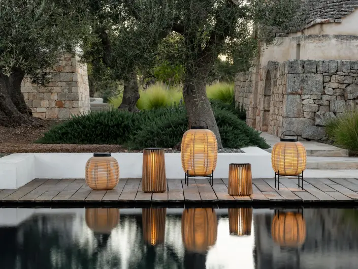 Tika and Maya solar lanterns by Vincent Sheppard, arranged on a wooden deck beside a pool, featuring handwoven rattan and teak designs with black handles and bases, softly illuminated.