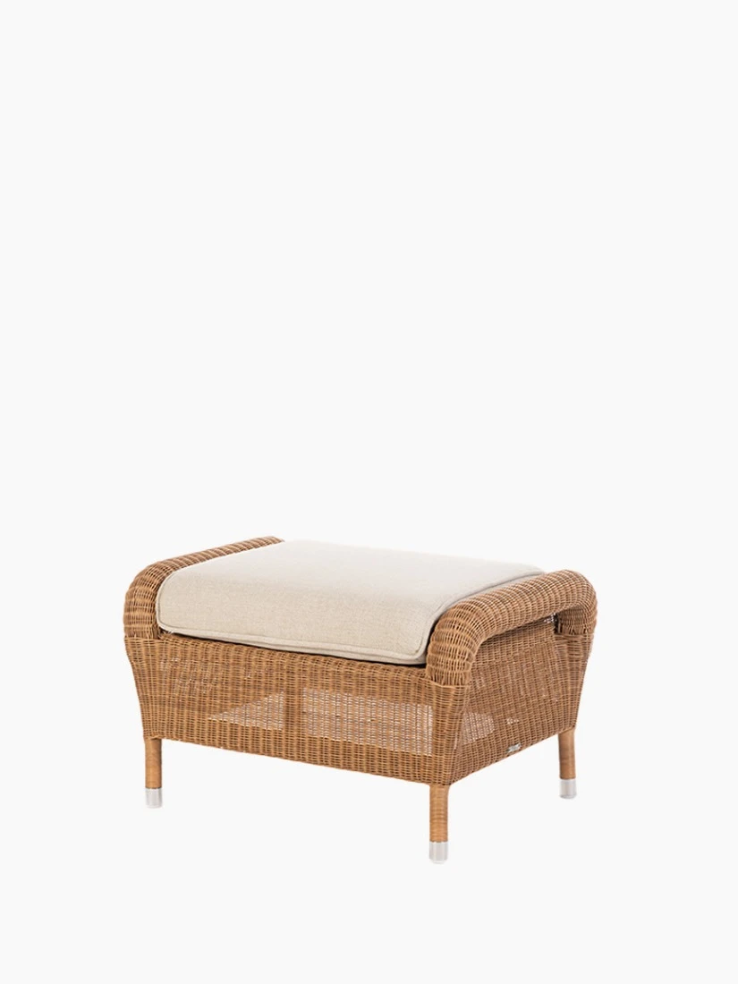 Angled view of Vincent Sheppard’s Riviera footrest in honey synthetic wicker with a beige cushion.