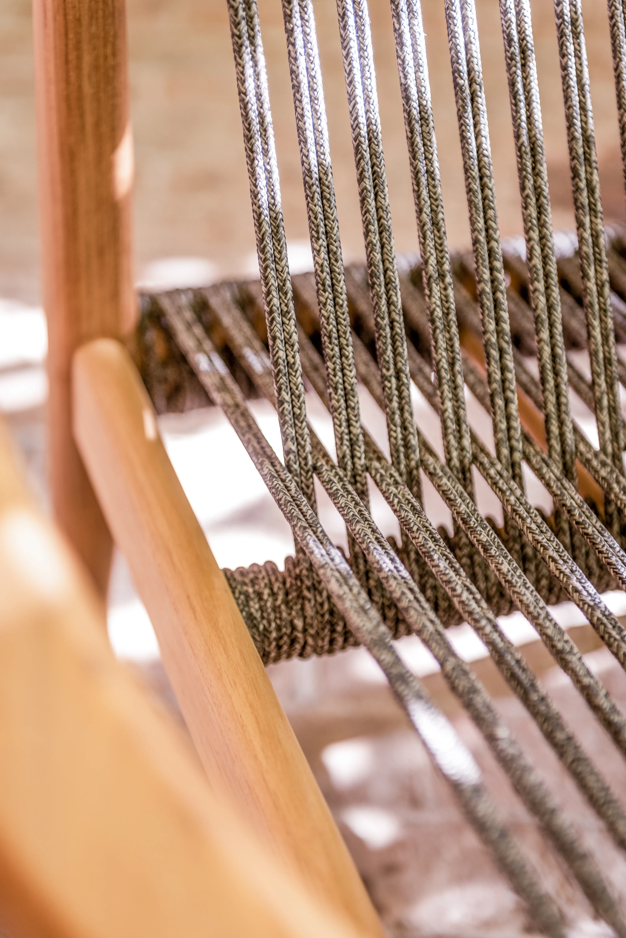 Close-up of a Vincent Sheppard chair featuring anthracite rope woven vertically across a warm teak wood frame. The braided rope adds texture and contrast to the smooth, natural finish of the teak, with sunlight enhancing the craftsmanship and material detail.