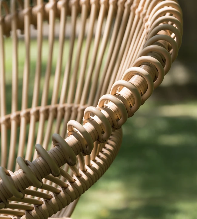 Detail of the Lazy Roy chair by Vincent Sheppard in camel synthetic wicker, highlighting its sculptural looped design against a blurred green outdoor background.