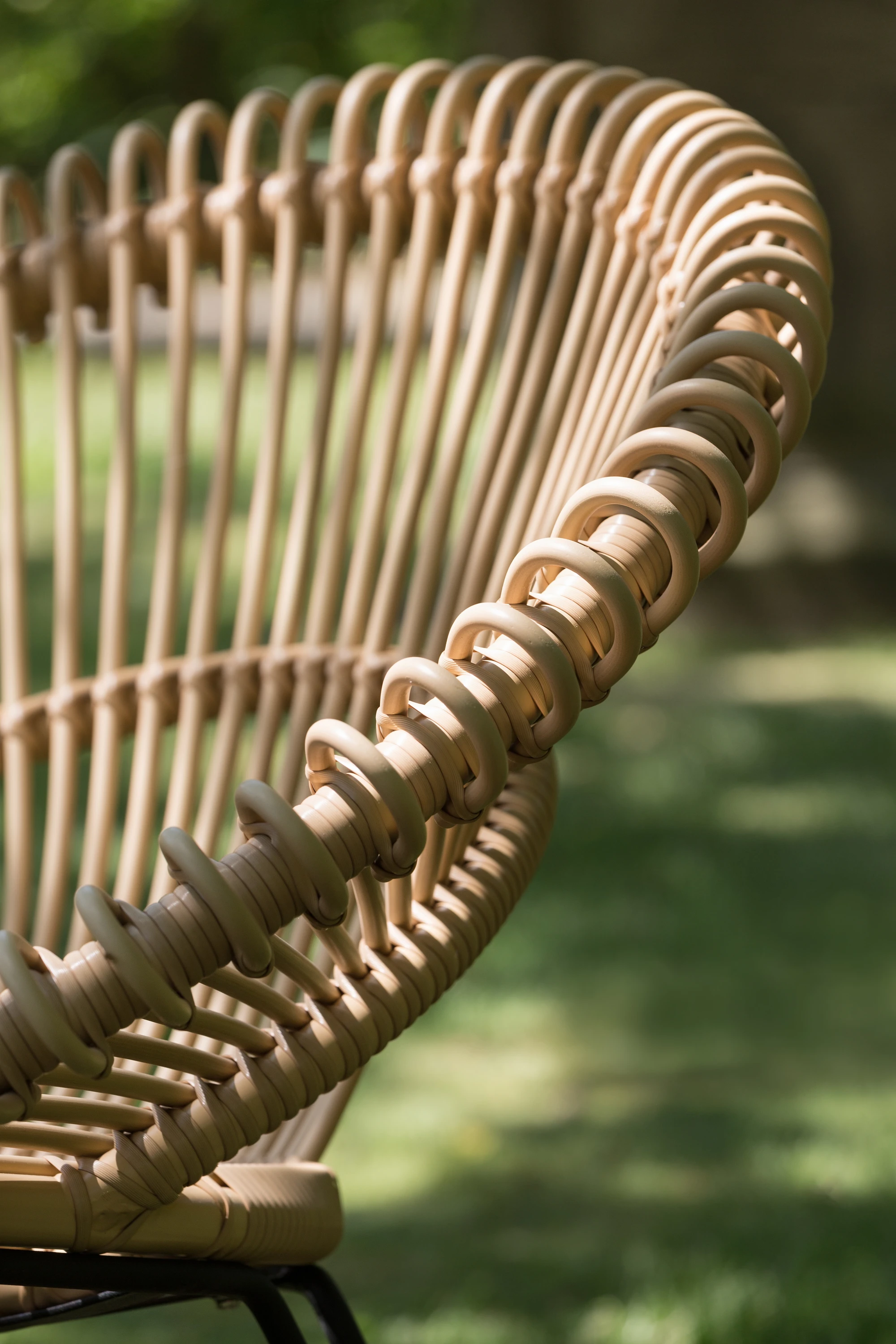 Detail of the Lazy Roy chair by Vincent Sheppard in camel synthetic wicker, highlighting its sculptural looped design against a blurred green outdoor background.