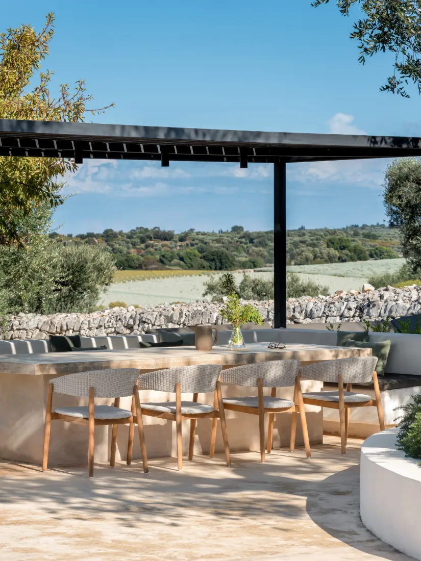 Mona dining chairs by Vincent Sheppard in Old Lace synthetic wicker with teak frames, arranged around a light stone outdoor table under a modern black pergola. 
