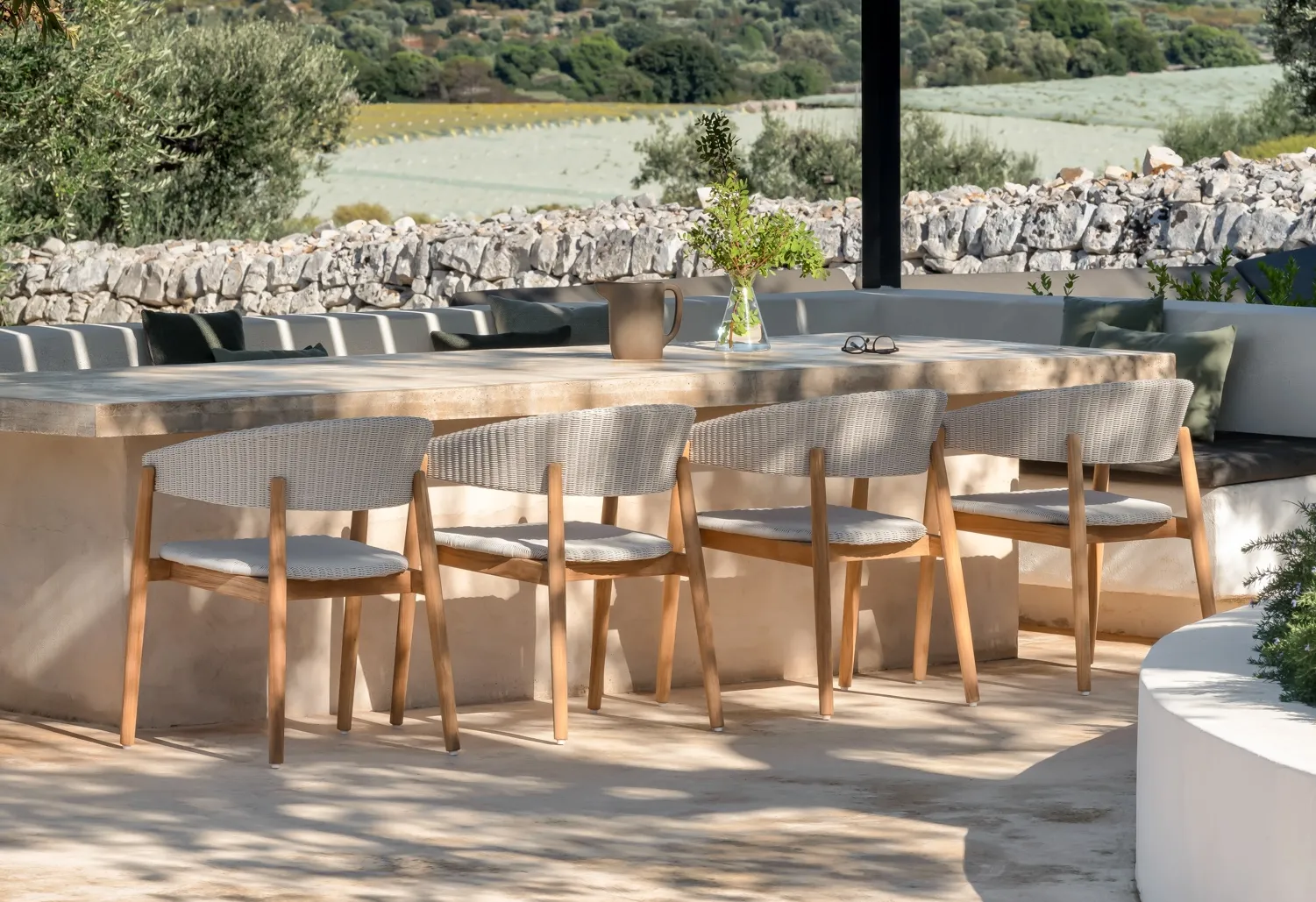 Mona dining chairs by Vincent Sheppard in Old Lace synthetic wicker with teak frames, arranged around a light stone outdoor table under a modern black pergola. 