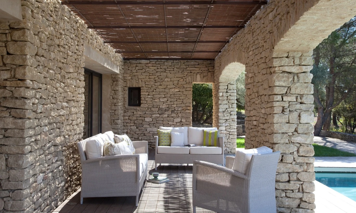 Safi lounge setting by Vincent Sheppard in Old Lace synthetic wicker, featuring a sofa and armchairs with white cushions, arranged on a shaded stone terrace beside a pool. The rustic stone walls and wooden pergola create a serene, Mediterranean-inspired atmosphere.