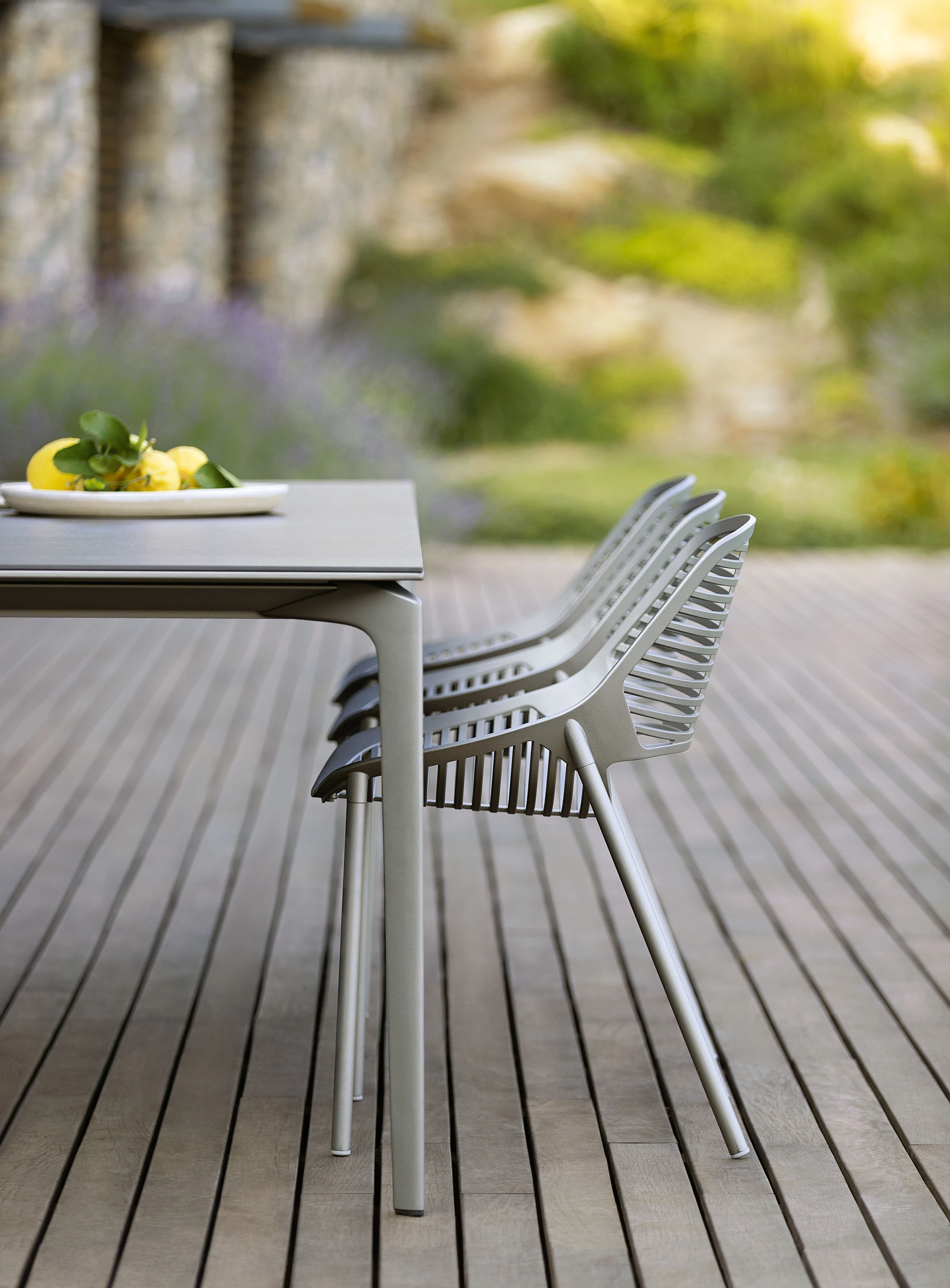 Allsize Table and Niwa chairs by FAST in  Iron Grey powder-coated aluminum, shown in an outdoor setting on a wooden deck with a natural garden backdrop.