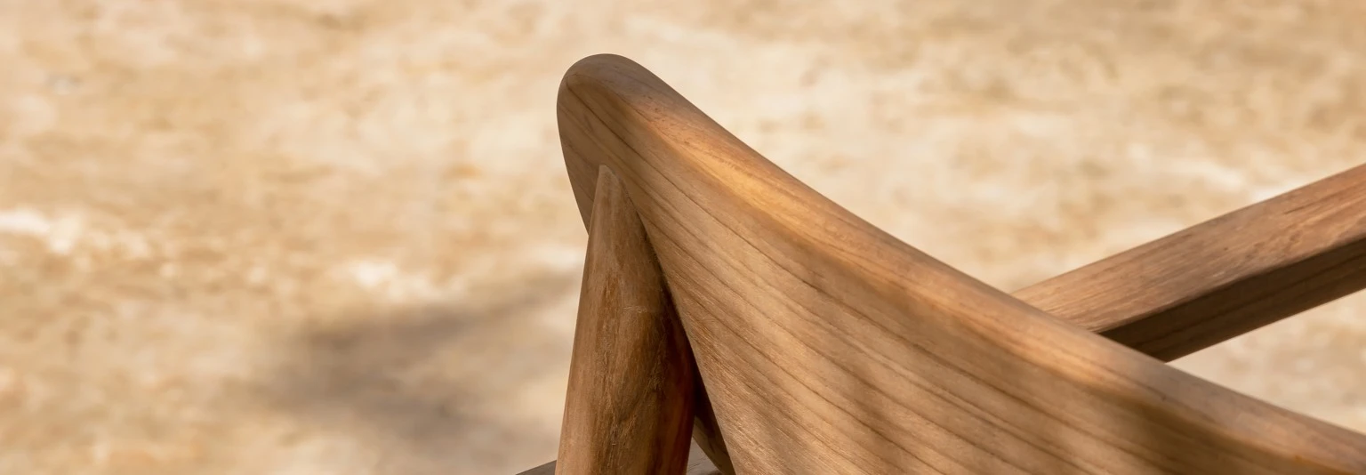 Freya Dining Chair by Vincent Sheppard, highlighting the smooth curves and joinery of its teak wood frame with a gently angled backrest and slatted seat, bathed in dappled sunlight